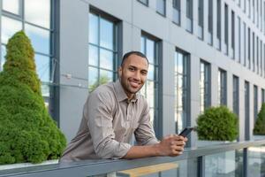 un joven latín americano empresario es en pie en el veranda de el oficina centro, utilizando un móvil foto