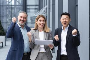 Businesswoman boss with her diverse team looking at the camera and rejoicing in success and victory, celebrating a successful contract a team of colleagues outside the office building with documents. photo