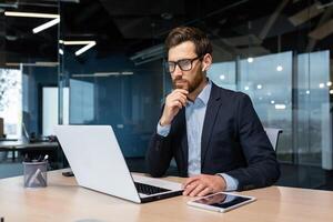 Senior businessman in glasses and beard is thinking about a decision, man is working inside the office and using laptop at work, mature investor in a business suit is sitting at the table photo