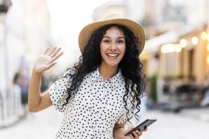 retrato de un contento joven indio mujer caminando en el ciudad en un soleado día, participación el teléfono en su mano, sonriente y saludo, ondulación a el cámara. foto