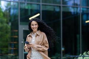 Young beautiful hispanic woman walking in the city, business woman holding phone in hands using smartphone app, woman smiling contentedly and happy outside office building with curly hair photo