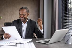 contento diverso masculino levantamiento mano con apretado puño mientras mirando a documento en mano por escritorio con ordenador personal. regocijado gerente leyendo información con aprobatorio resultado de salario creciente pedido. foto