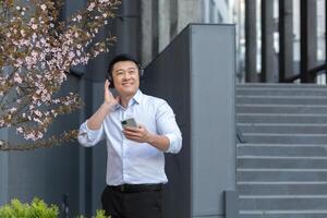 satisfecho joven hermoso asiático hombre en blanco camisa sostiene teléfono y escucha a audio libro, podcast en negro auriculares. caminando en el calle cerca un moderno edificio y un hermosa magnolia árbol. foto