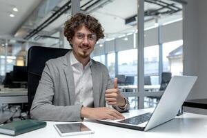 Professional young male office worker with curly hair showing approval with thumbs up gesture at his workplace in a corporate office. photo