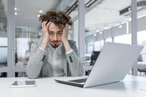 un joven empresario con Rizado pelo aparece estresado y cansado mientras trabajando en un ordenador portátil en un moderno oficina. foto