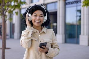 Portrait of young beautiful African American woman, student smiling and looking at camera holding phone in hands using headphones for listening to music and online radio with podcasts. photo