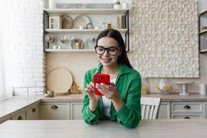 hermosa mujer en lentes y verde camisa utilizando rojo móvil teléfono a hogar foto