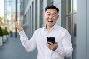 retrato de un contento joven asiático hombre en un blanco camisa en pie fuera de un edificio, participación un teléfono, mirando sorprendido y sonriente a el cámara, disfrutando su éxito. foto