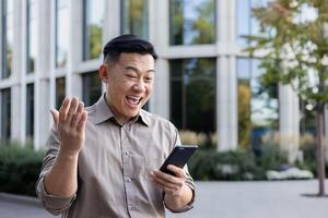 contento joven asiático hombre en pie cerca oficina edificio y leyendo mensaje en teléfono, contento a recibir genial Noticias y éxito. foto