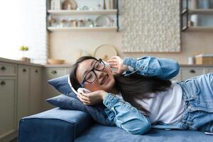 de cerca foto de joven mujer acostado en sofá a hogar escuchando a música utilizando auriculares, sonriente y relajante en cocina