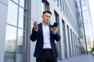 Serious young Asian businessman standing outside office building, holding phone, looking frustrated and worried at screen with arms outstretched. photo