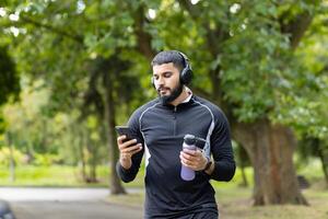 A fit man in sportswear takes a break in a lush park to use his smartphone and drink water, embodying an active, healthy lifestyle. photo