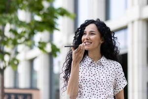 sonriente mujer en lunares blusa utilizando voz asistente en teléfono en urbano configuración, transporte facilitar y moderno tecnología. foto