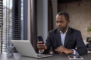 Focused African American businessman multitasking with a smartphone and laptop in a modern office setting. photo