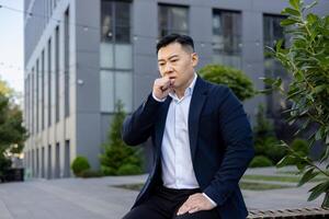 A young Asian man is not feeling well, he is sitting sick outside his office on a bench and coughing covering his mouth with his hand. photo