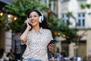 hermosa joven latín americano mujer camina en el noche ciudad en un viaje, mujer con auriculares escucha a música y bailes y canta, usos un en línea radio solicitud en su teléfono foto