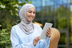 Professional happy Muslim woman with hijab holding a tablet, enjoying technology outside a modern office setting. photo