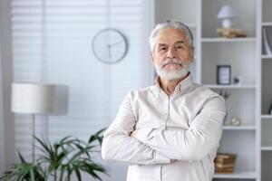 un retrato de un confidente mayor hombre con doblada brazos en pie en un elegantemente amueblado hogar interior demostración contentamiento y madurez. foto