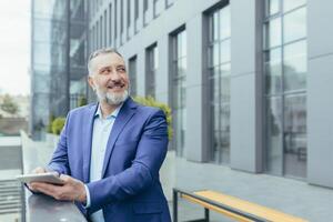 Senior handsome gray-haired male teacher, professor, lecturer is standing in a suit on the balcony in a modern campus, holding a tablet in his hands. He looks up at someone, smiles, talks. photo