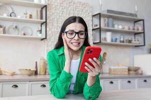 Shocked young beautiful woman in glasses reading message from phone, teenager in kitchen. photo