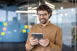 Smiling man in spectacles holding digital tablet while standing in corporate building with glassy walls. Confident gadget user checking options and possibilities for work of brand new device. photo