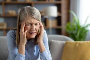 A mature woman sits in her living room, visibly distressed with a severe headache, holding her temples in pain and discomfort. photo