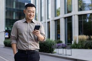 Asian young man businessman, office worker, manager standing outside office center and using mobile phone. photo