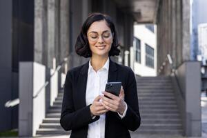 Close-up photo of a smiling young business woman in a suit and glasses standing outside an office building and using a mobile phone.