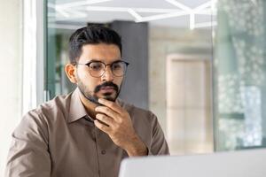 Young Indian businessman in a serious thinking pose, working on his laptop at an office, showing dedication and focus. photo