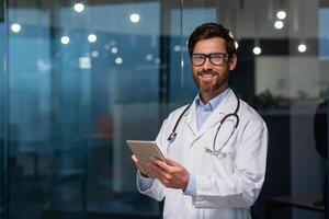 retrato de maduro y experimentado doctor, hombre en médico Saco y lentes mirando a cámara, Doc utilizando tableta computadora, trabajando dentro moderno oficina clínica foto