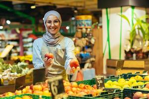 retrato de vendedora en supermercado, contento mujer en hijab sonriente y mirando a cámara, vendedor participación manzanas en vegetal sección, musulmán mujer en lentes y delantal entre estantería con bienes foto