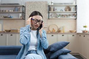 joven enfermo mujer a hogar tiene un grave dolor de cabeza, asiático mujer sentado en el sofá en el cocina hablando en el teléfono, vocación el médico para ayuda, y consultante foto