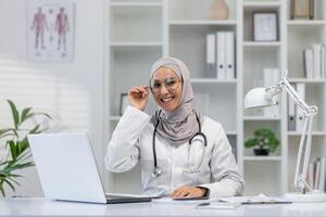 A cheerful, professional doctor wearing a hijab smiles warmly in her well-equipped clinic office, adorned with medical books and a healthy plant, projecting confidence and compassion amid a modern medical setting. photo