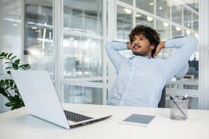 A young man with curly hair sits back, hands behind head, reflecting or taking a break in a well-lit office environment. The scene evokes a sense of calm and contemplation. photo