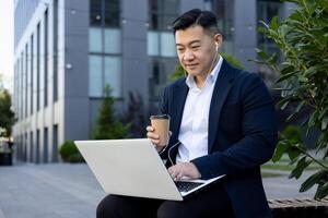 asiático joven masculino empresario sentado en un banco cerca un oficina edificio vistiendo auriculares, participación un taza de café en su mano y utilizando un ordenador portátil. foto