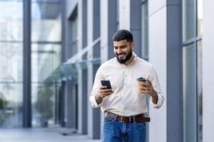 A cheerful bearded young adult is engrossed in his smartphone while casually holding a takeaway coffee cup. He's strolling in front of a modern glass building, exemplifying a relaxed yet dynamic urban lifestyle. photo