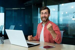 Portrait of successful mature businessman in red shirt at workplace inside office, man celebrating victory and successful achievement results at work, looking at camera, holding hand up. photo