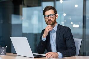 Mature serious thinking businessman working inside office, boss investor in business suit working at table with laptop brainstorming business strategy and plan. photo