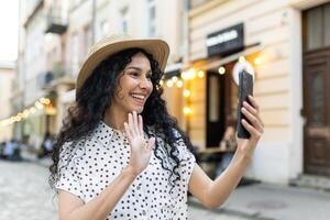 un atractivo indio turista mujer es felizmente caminando alrededor el ciudad haciendo un llamada a su parientes en el teléfono, atentamente sonriente y saludo por ondulación su mano a el cámara. foto