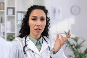 Female doctor is serious, looking worriedly into smartphone camera talking to patient remotely, reporting disappointing results online, Hispanic woman in white medical coat using app on phone call. photo