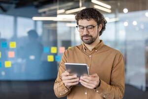 A focused business professional stands in a modern office, using a tablet. His casual business attire and the office's glass background with sticky notes add to the corporate atmosphere. photo