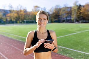 joven mujer atleta corredor en pie en estadio con teléfono y auriculares y melodías a escucha a música, audio libro, podcast. sonriente a el cámara. foto