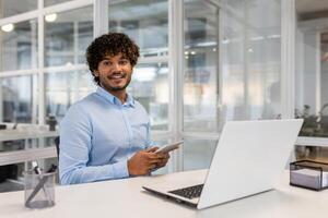 un joven profesional hombre sentado a un escritorio en un brillante oficina, trabajando en un ordenador portátil mientras participación un teléfono inteligente, exudando un confidente y simpático comportamiento. foto