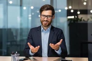 retrato de un simpático joven hermoso hombre sentado en el oficina a el mesa vistiendo auriculares y hablando a el cámara. negocio reunión, tutor apoyo, trabajo entrevista, consulta foto