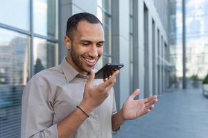 sonriente Hispano empresario grabación voz mensaje, contento hombre fuera de oficina edificio leyendo comandos en teléfono inteligente para ai buscar foto