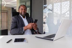 retrato de mayor gris peludo africano americano jefe, hombre sonriente y mirando a cámara a lugar de trabajo dentro oficina, empresario participación teléfono, utilizando en línea aplicación en teléfono inteligente foto