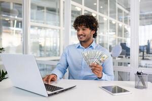 A cheerful young man in a blue shirt holds US dollar bills while sitting at a desk with a laptop in a bright office setting, showcasing financial success. photo