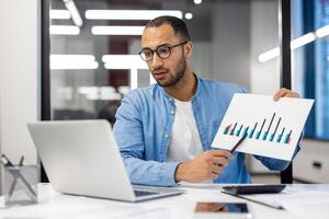 A young Latin American man works in the office, sits at a table in glasses, shows documents with graphs on a laptop camera, conducts an online meeting. photo