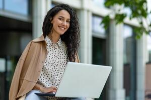 A smiling Hispanic woman uses her laptop while seated outdoors. Dressed in a stylish trench coat, she works amidst modern urban settings. photo