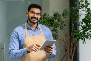 alegre masculino pequeño negocio propietario en delantal opera tableta en verde lleno de plantas contemporáneo espacio de trabajo, ejemplificando moderno emprendimiento foto